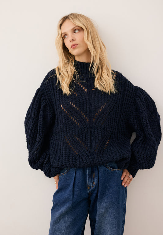 Modelka patrząca w bok ubrana w ażurowy sweter ROSSO w kolorze granatowym i ciemne jeansy LEYA.