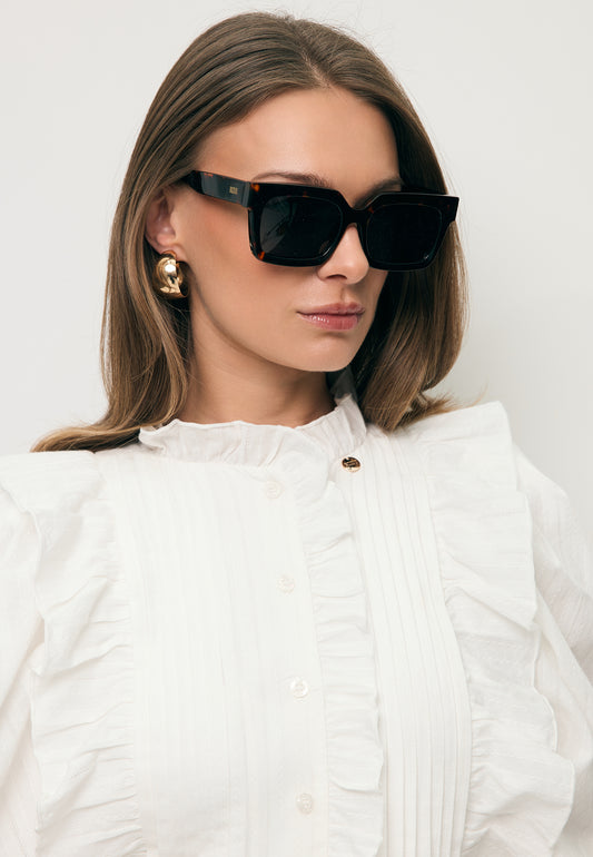 Modelka w białej koszuli i okularach przeciwsłonecznych AVOLA brązowych.