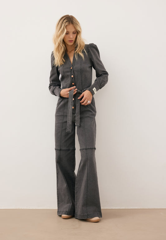 Ciemnoszary jeansowy kombinezon z długimi rękawami, szerokimi nogawkami i guzikami z przodu. W talii przewiązany paskiem. Osoba stoi na chodniku przed kamiennym budynkiem.