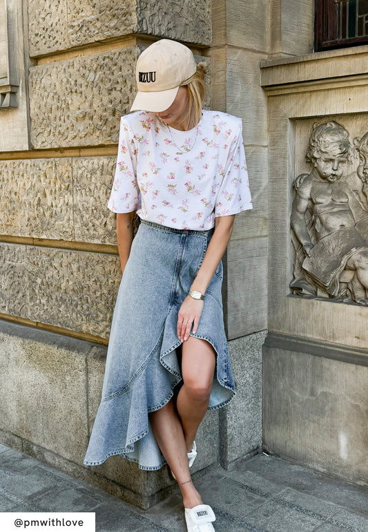 Modelka ubrana w białą koszulkę z kwiatowym nadrukiem, beżową czapkę z daszkiem oraz jeansową spódnicę z falbaną i asymetrycznym dołem.