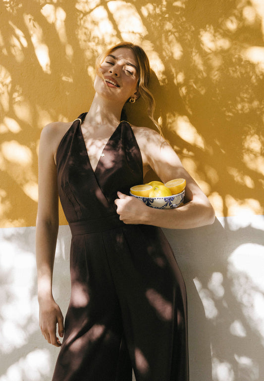 Modelka oparta o żółta ścianę trzyma w ręce miskę z cytrynami i ubrana jest w brązowy kombinezon ARATIKA.