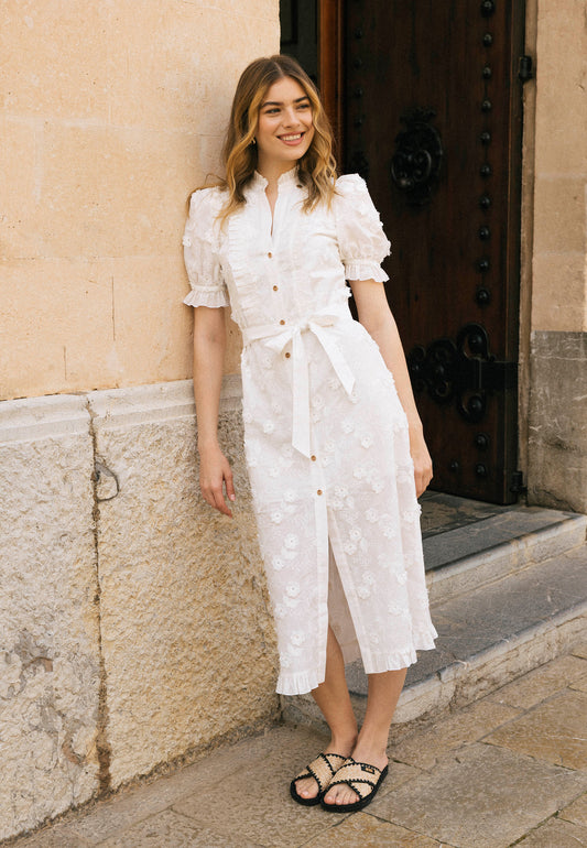 Modelka, ubrana w białą midi sukienkę typu szmizjerka NATTA, opiera się o ścianę.