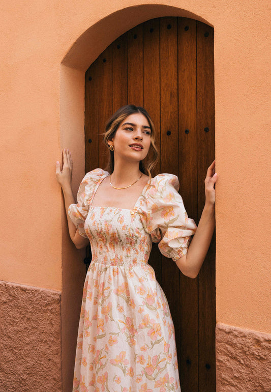 Modelka w kwiecistej sukience LUMIA stoi w łuku drewnianych drzwi, oparta o framugę.