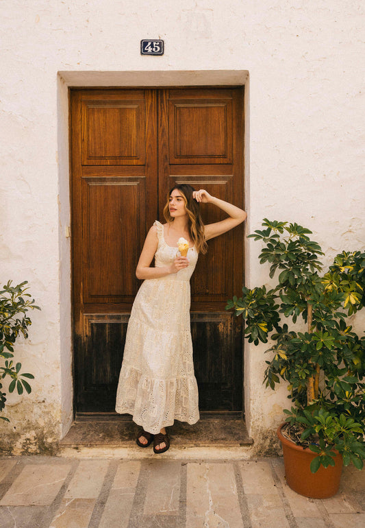Kobieta w jasnej, koronkowej sukience ROCHELLE stoi z lodem przed drewnianymi drzwiami, otoczona roślinami w doniczkach.
