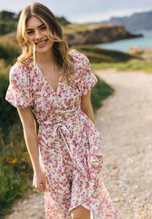 Uśmiechnięta kobieta w kwiecistej sukience kopertowej z krótkim rękawem ISLAY idzie ścieżką nadmorską, w tle woda i skały.