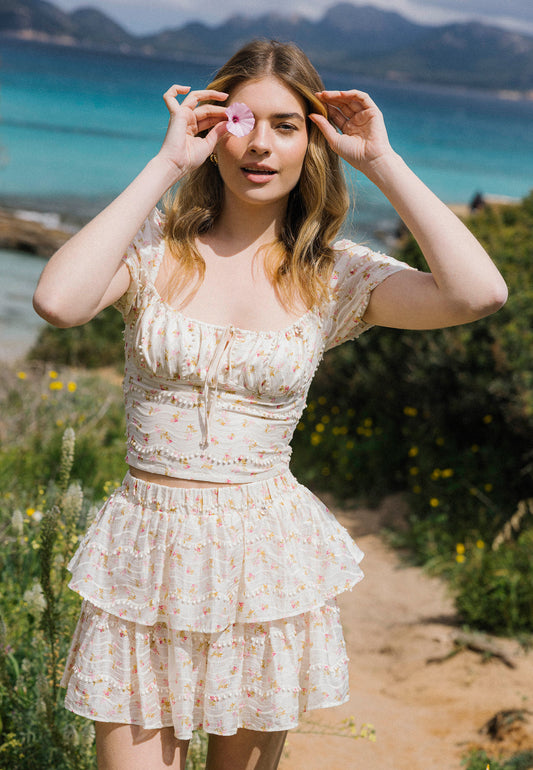 Uśmiechnięta kobieta w romantycznym komplecie z falbanami VANNES i COKO pozuje na ścieżce wśród dzikich kwiatów, trzymając różowy kwiatek przy oku. W tle błękitna laguna i górzysty krajobraz.