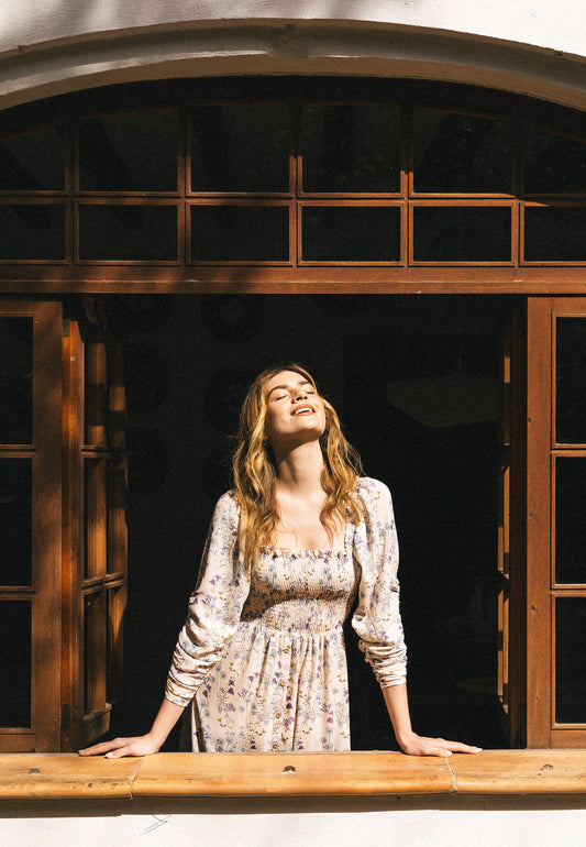 Modelka ubrana w długą sukienkę PATRYA w delikatny, kwiatowy wzór wychyla się przez drewniane okno, twarz zwrócona ku słońcu z zamkniętymi oczami, w świetle porannego blasku.