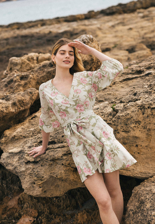 Kobieta w kremowej sukience mini AMBROSIA z różowym kwiatowym nadrukiem i rękawem 3/4, oparta o skały, osłania oczy przed słońcem.