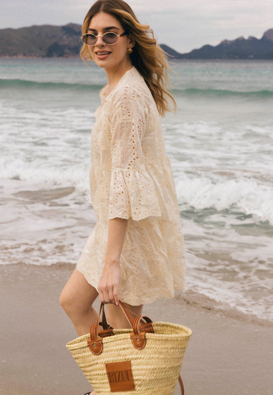 Kobieta w kremowej sukience ażurowej SANTIA z rękawami 3/4 spaceruje brzegiem morza, trzymając pleciony koszyk z brązowymi rączkami i logo BIZUU.