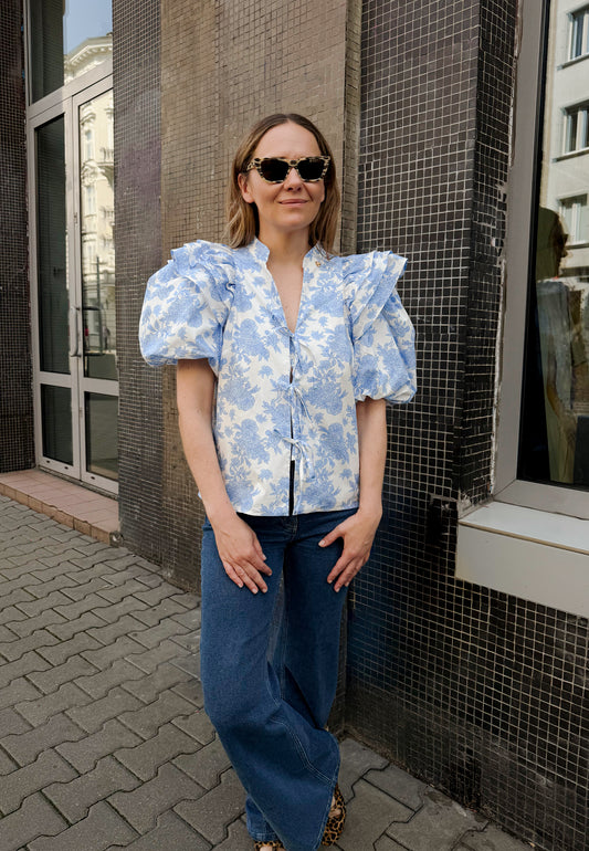 Modelka stojąca przed budynkiem ubrana w bluzkę ALIANNA w kwiatowy print w kolorze błękitu z wiązaniami z przodu i spodnie ATLANTA.