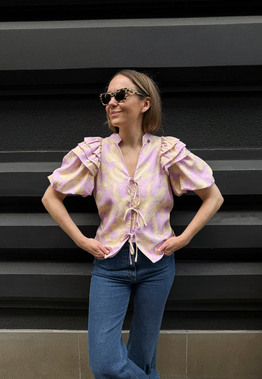 Modelka na tle budynku ubrana w bawełnianą bluzkę ALIANNA wiązaną na przodzie oraz w granatowe jeansy ATLANTA.