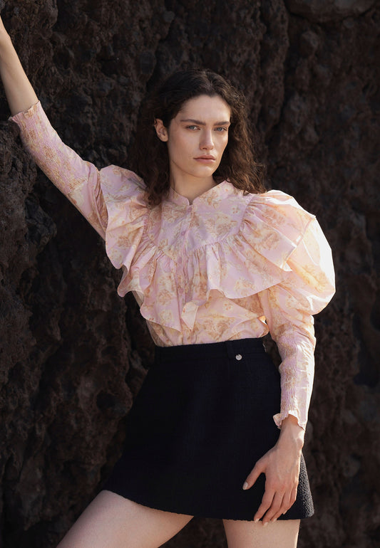 Modelka oparta o skałę ubrana w różową koszulę z ozdobną falbaną ANTUANA oraz czarną tweedową spódniczkę BREDA.