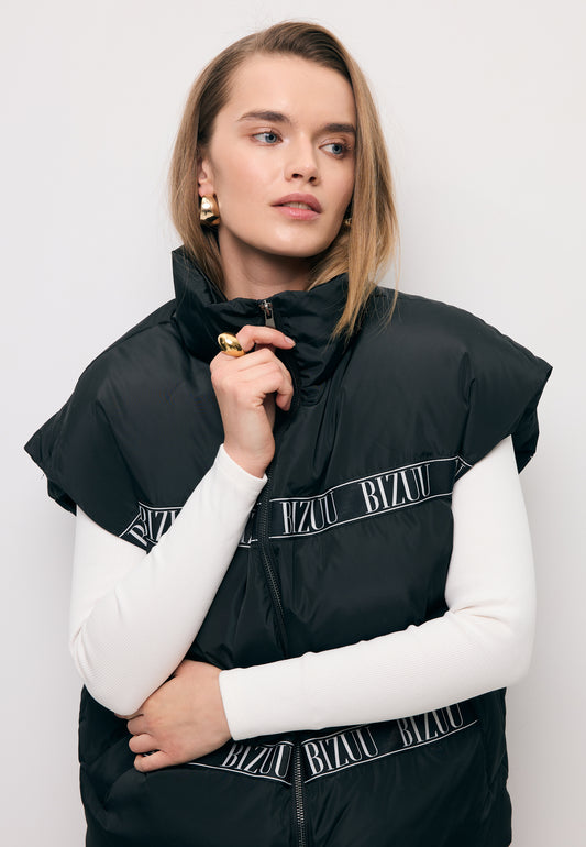 Zbliżenie na modelkę zapinającą czarną puchową kamizelkę z logowaną taśmą AYASE.