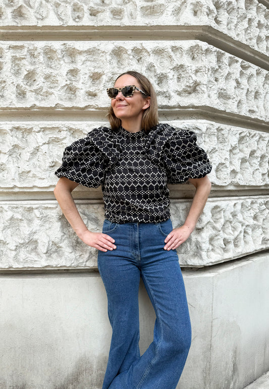 Modelka oparta o ścianę budynku z rękoma w kieszeniach spodni jeansowych ATLANTA. Ubrana w czarną, bawełniana bluzkę CASSANDRA z kontrastowym haftem.