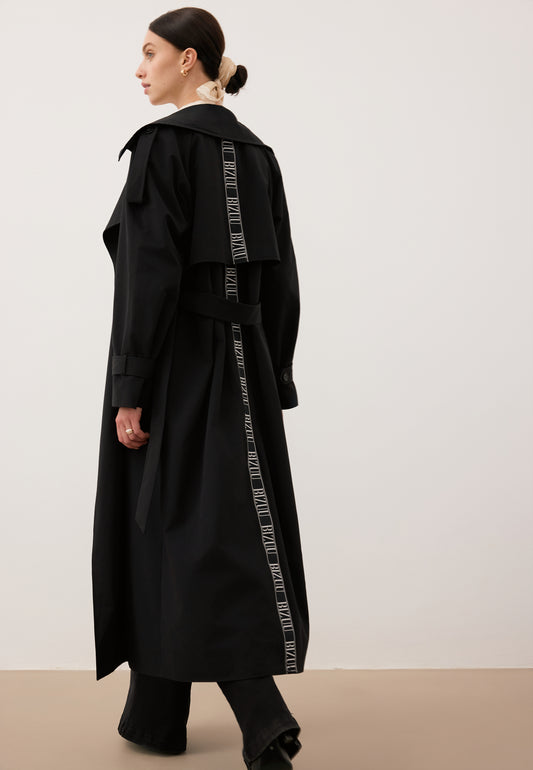 Modelka na tle białej ściany robiąca krok w przód ubrana w czarny płaszcz RACHELLA z logowaną taśmą na plecach.