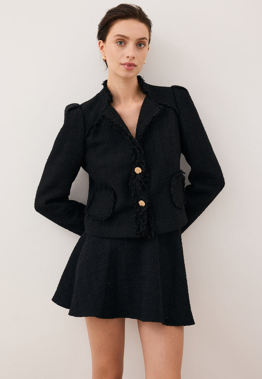 Modelka oparta o ścianę z rękoma z tyłu ubrana w mini spódniczkę WENDYS i czarną marynarkę z logowanymi guzikami DITAS.