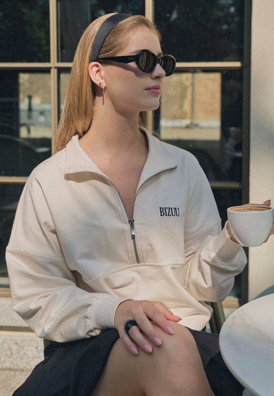 Kobieta w czarnych okularach przeciwsłonecznych BIZUU ENNA, siedząca przy stoliku z filiżanką kawy. Ma na sobie jasną bluzę z suwakiem i logo BIZUU, nosi czarną opaskę i złote kolczyki – elegancki look inspirowany stylem lat 90.