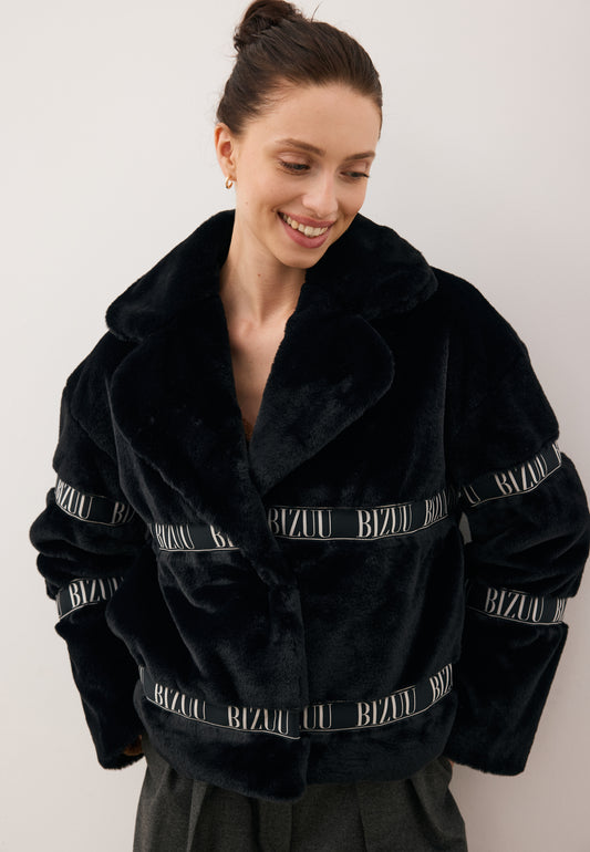 Modelka siedząca na krześle ubrana w pudełkowe futerko GREGORIA czarne z logowaną taśmą.