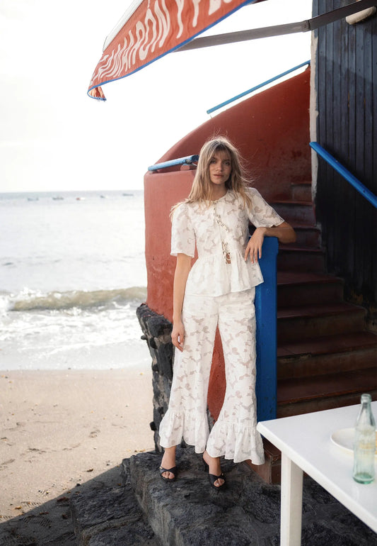 Modelka oparta o murek ubrana w kremowy zestaw we wzór kwiatowy - bluzka SAVENNA i spodnie LAVENDOU.