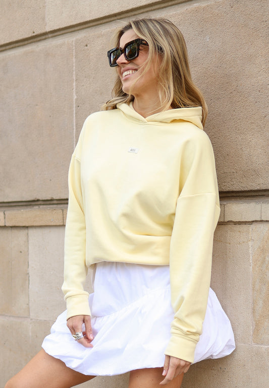 Zbliżenie na modelkę opartą o ścianę ubraną w żółtą bluzę LINDA i białą mini spódniczkę LILIBET.