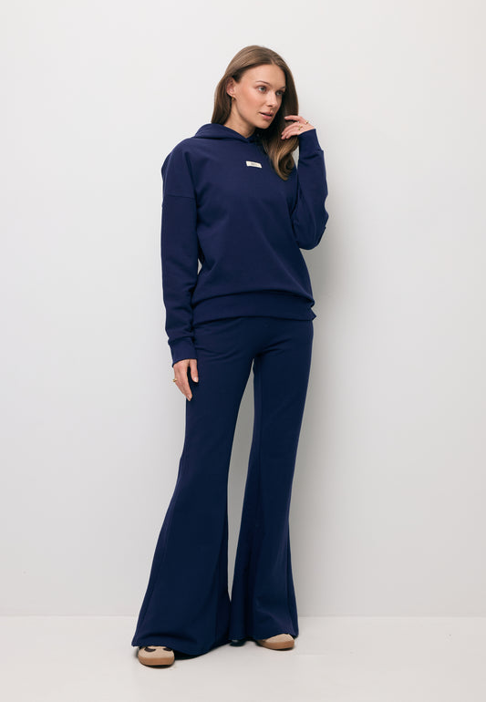Modelka na tle białej ściany ubrana w granatowy komplet dresowy - bluza z kapturem LINDI oraz spodnie typu flare TEIA.