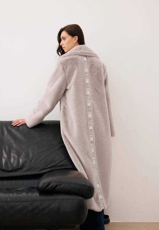 Modelka stojąca tyłem oparta o kanapę, ubrana w długie, beżowe futro LIVORNO z lampasem na plecach.