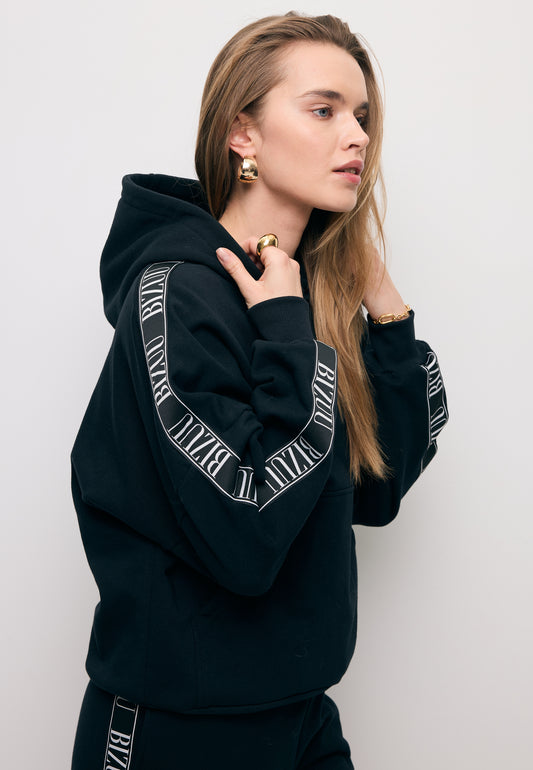 Zbliżenie na modelkę stojącą bokiem w czarnej bluzie z kapturem i lampasem na rękawie MAYRA.