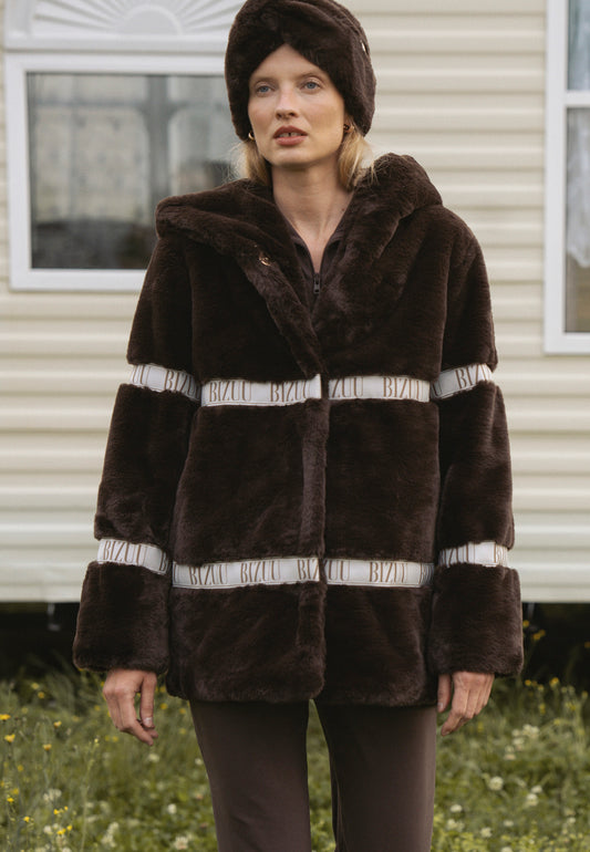Modelka na polanie na tle kampera w brązowym futrze z kapturem i logowaną taśmą MILANO.