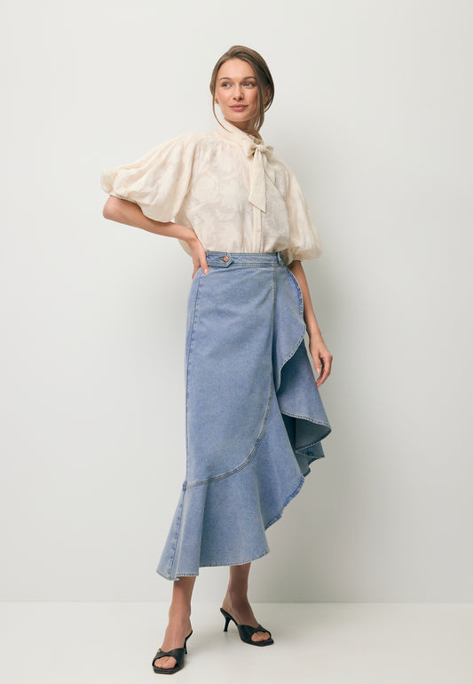 Modelka stojąca po skosie ubrana w jeansową spódniczkę z ozdobną falbaną NIIA oraz koszulą w kwiatowy wzór AZALEA w kolorze kremowym.