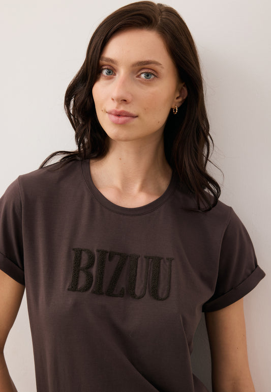 Zbliżenie na modelkę ubraną w czekoladowy T-shirt NIKKO z brązowym logo na przodzie.
