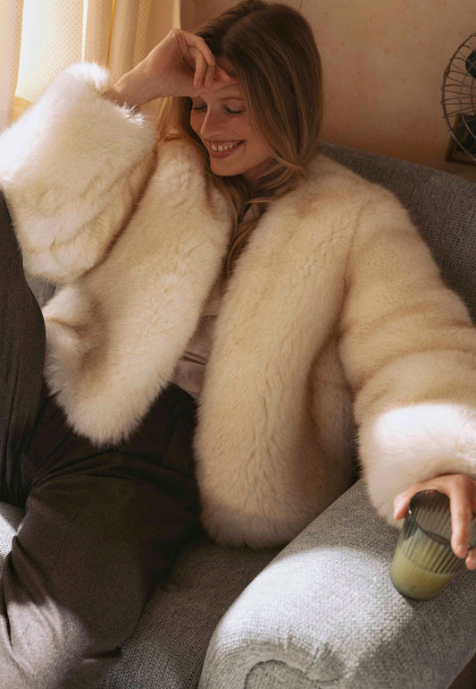 Śmiejąca się modelka siedzi na kanapie w kremowym, krótkim futerku NOVARA.
