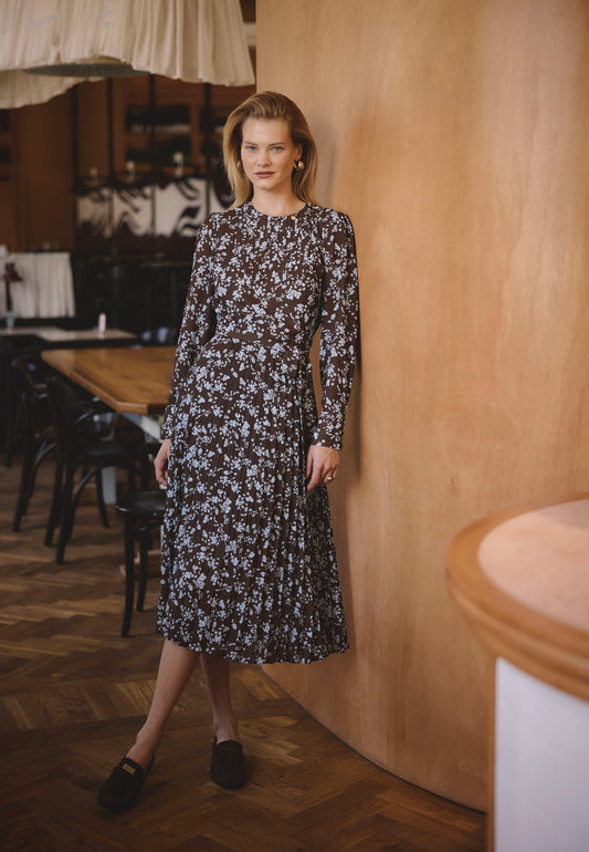 Modelka ubrana w brązową sukienkę w niebieskie kwiatki NYLA oparta o drewnianą ścianę.