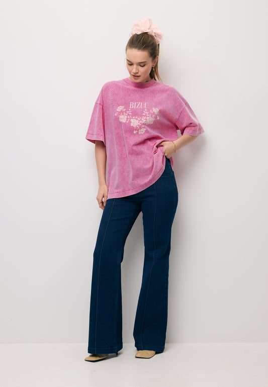 Modelka na tle białej ściany z ręką ugiętą na biodrze ubrana w różowy T-shirt oversize PAPLYS oraz granatowe jeansy typu flare BELINNA.