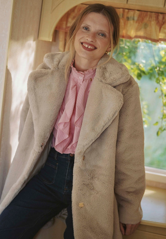 Modelka siedząca na blacie ubrana w beżowe futro ROMA, różową koszulę SANNI i jeansy DELPHI.