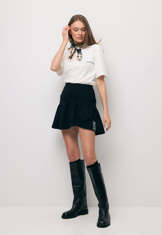 Modelka na tle białej ściany ubrana w T-shirt kremowy z ozdobnym logo i poduszkami MELISA oraz mini spódniczkę w kolorze czarnym TULIAN.