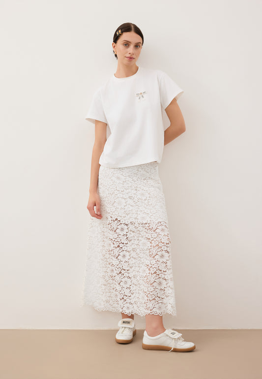 Modelka oparta o ścianę z jedną ręką zgiętą do tyłu ubrana w T-shirt z logowaną kokardką TISHALLE, białą, koronkową spódniczkę MILKY i buty MOBBY.