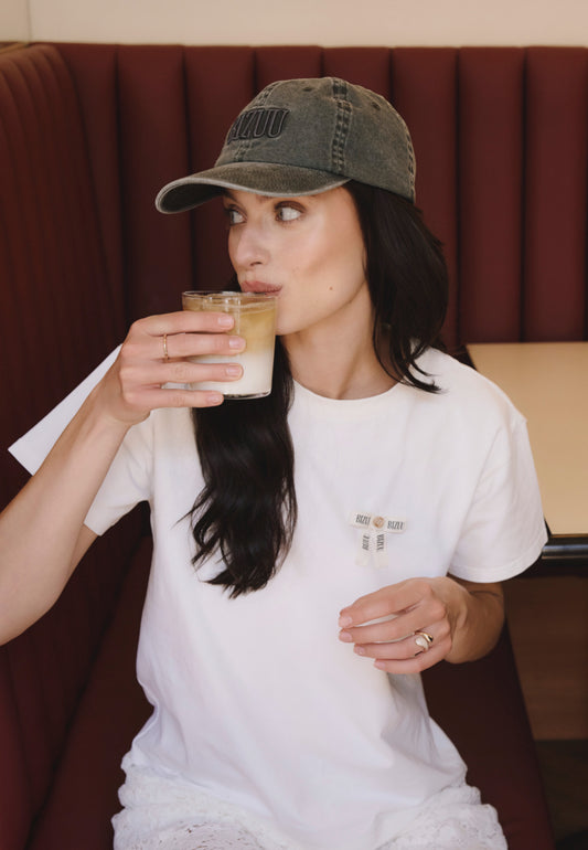 Modelka siedząca w kawiarni i pijąca kawę, ubrana w szarą czapkę z daszkiem ZANE oraz biały T-shirt z logowaną kokardką TISHALLE.