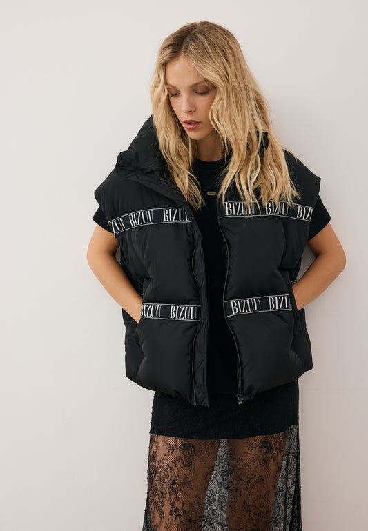 Modelka z rękoma w kieszeniach czarnej, puchowej kamizelki AYASE z taśmą z logo, dodatkowo ubraną ma koronkową spódniczkę FLACE.