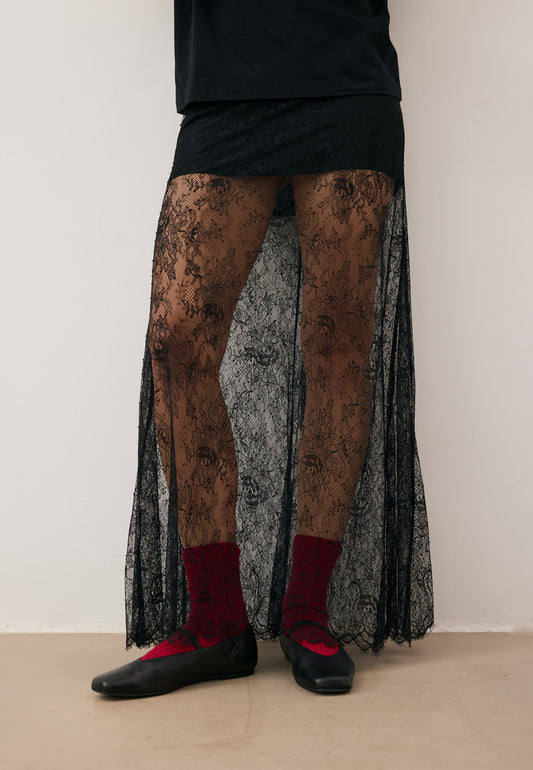 Modelka z jedną ręką na biodrze ubrana w czarną, koronkową spódnicę FLACE, czarny, bawełniany T-shirt TISHAL i okulary przeciwsłoneczne.