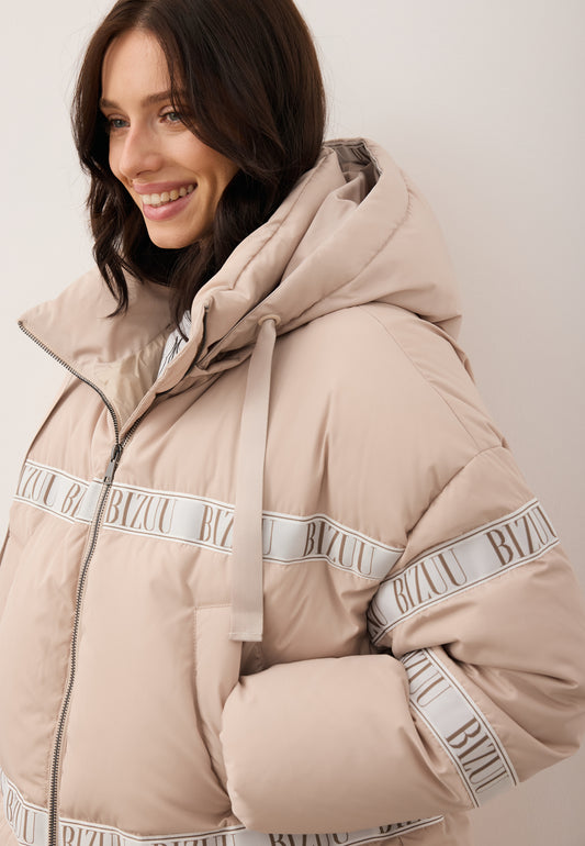 Zbliżenie na uśmiechniętą modelkę trzymającą rękę w kieszeni ubrana w beżową kurtkę z naturalnym wypełnieniem TOKYO z ozdobnym lampasem i kapturem.