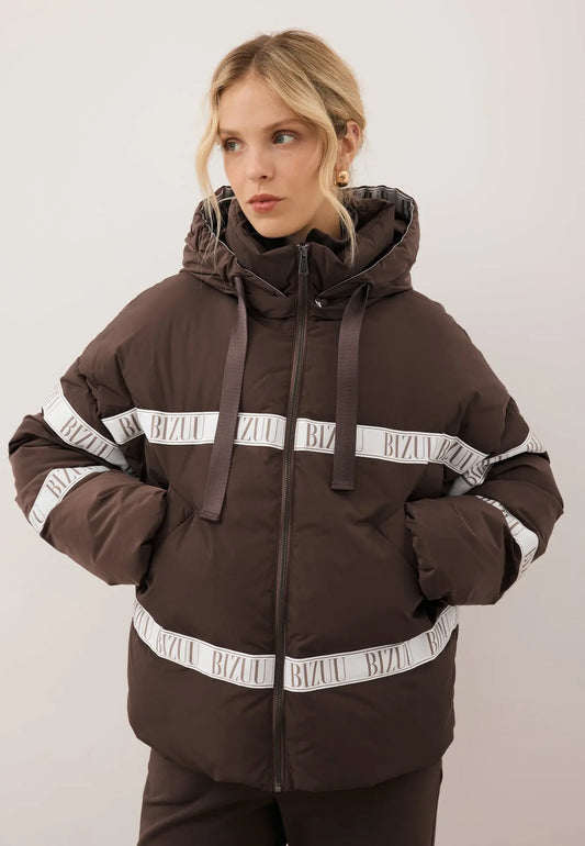 Zbliżenie na modelkę z rękoma w kieszeni brązowej kurtki TOKYO z naturalnym wypełnieniem oraz logo.
