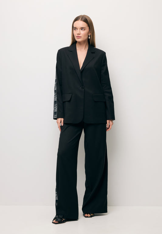 Modelka na tle białej ściany ubrana w czarny garnitur z białym lampasem - marynarka VANDA i spodnie MICHAEL.
