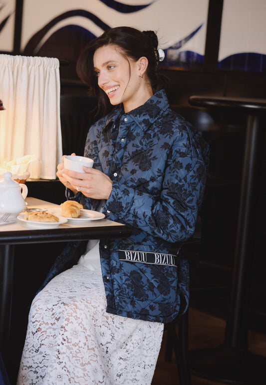 Modelka siedząca w kawiarni z filiżanką kawy w ręce, ubrana w białą koronkową spódniczką MILKY i kurtkę pikowaną w print MARIN.