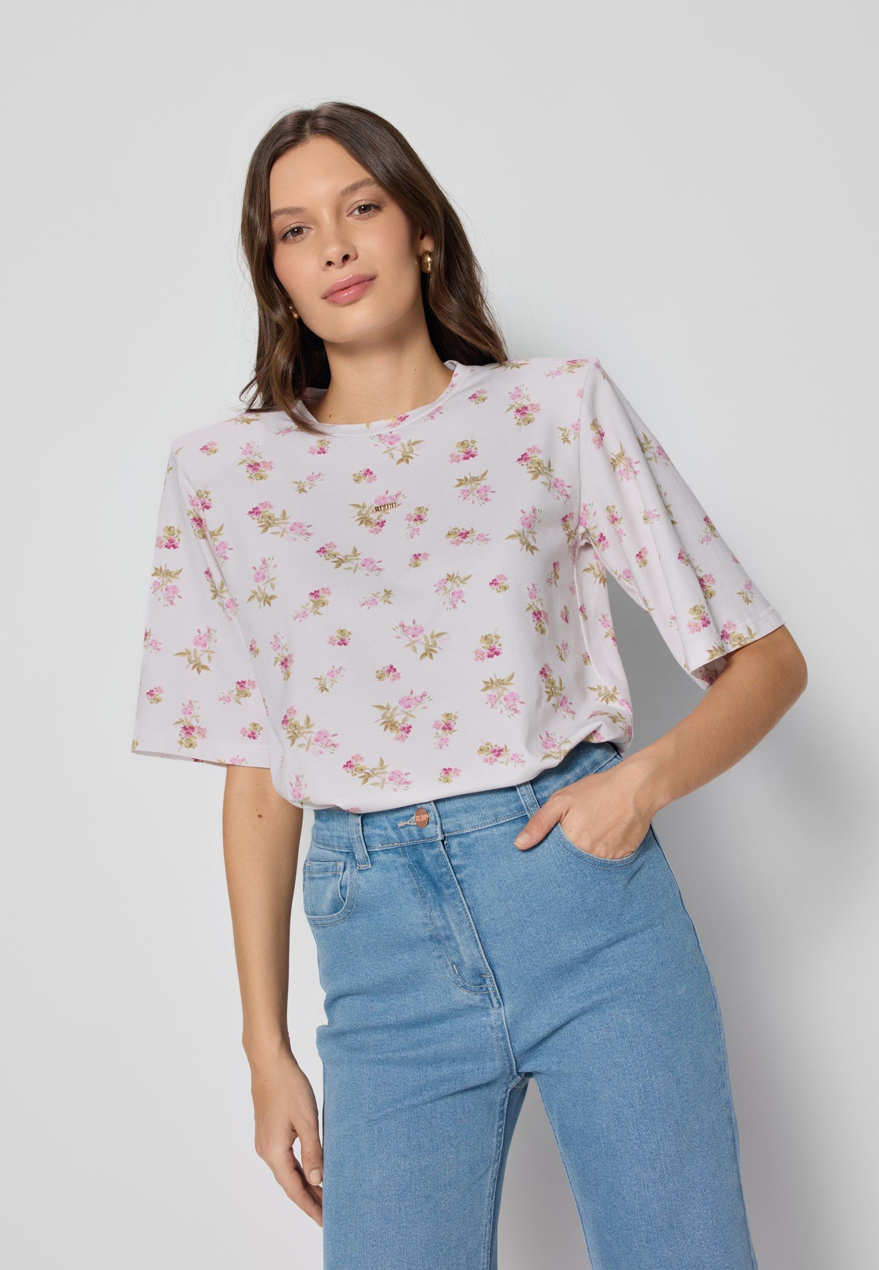 Niebieskie dżinsy noszone z białym t-shirtem w kwiaty, osoba stoi na neutralnym tle z ręką w kieszeni.