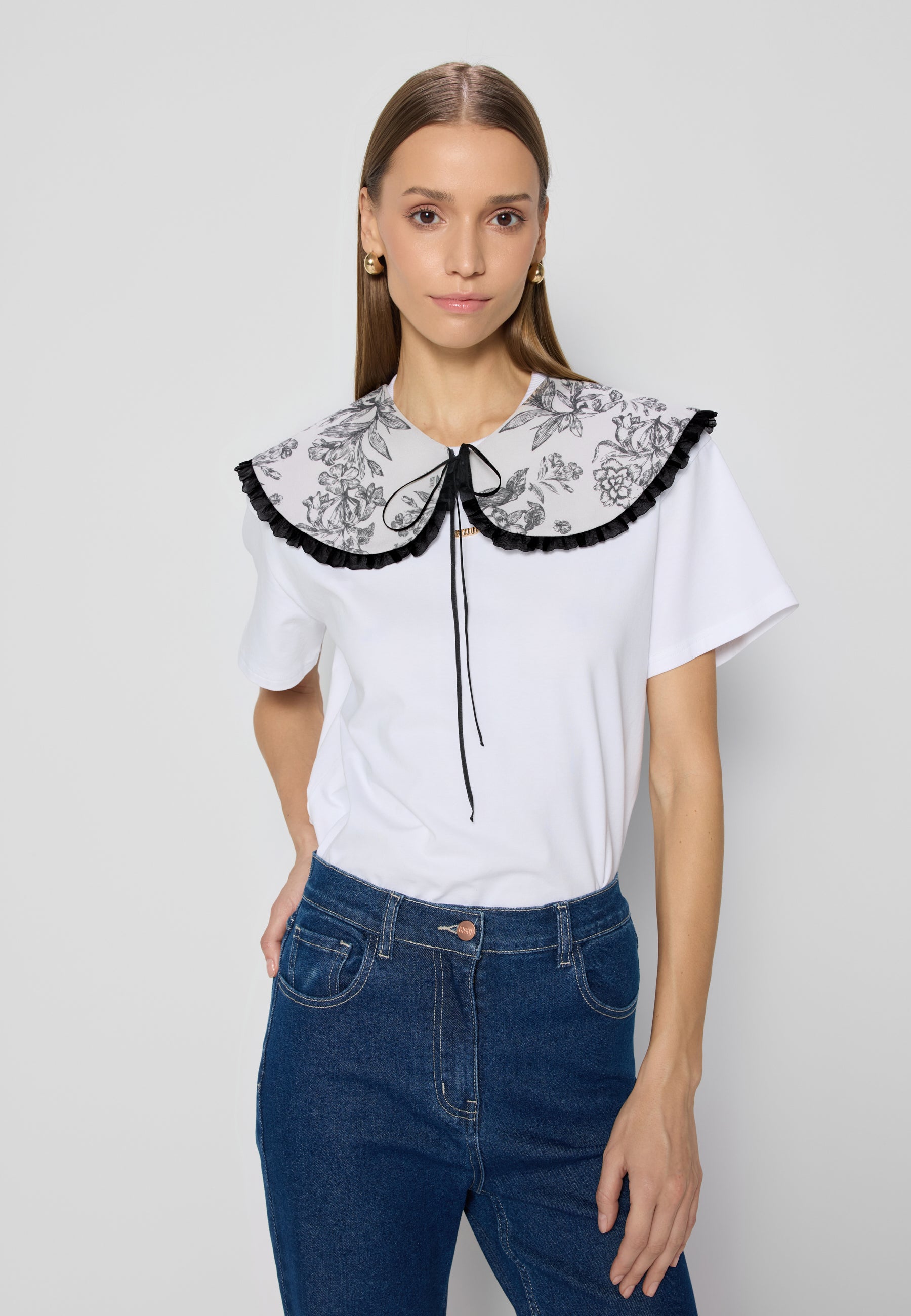 Niebieskie jeansowe spodnie dzwony noszone z białym t-shirtem z dużym kołnierzem w kwiaty i czarną lamówką. Osoba stoi na jasnym tle z ręką w kieszeni.