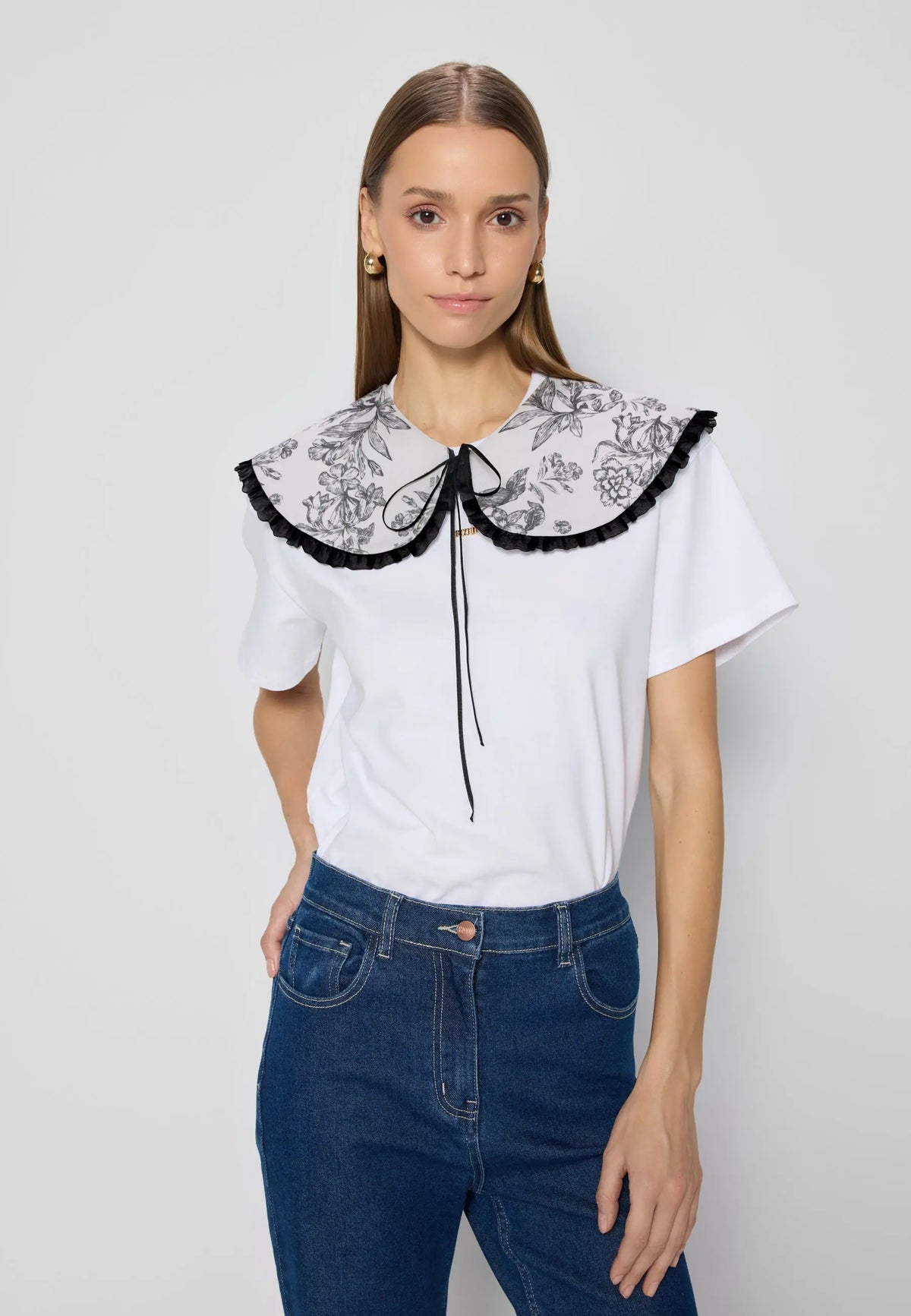 Niebieskie jeansowe spodnie dzwony noszone z białym t-shirtem z dużym kołnierzem w kwiaty i czarną lamówką. Osoba stoi na jasnym tle z ręką w kieszeni.