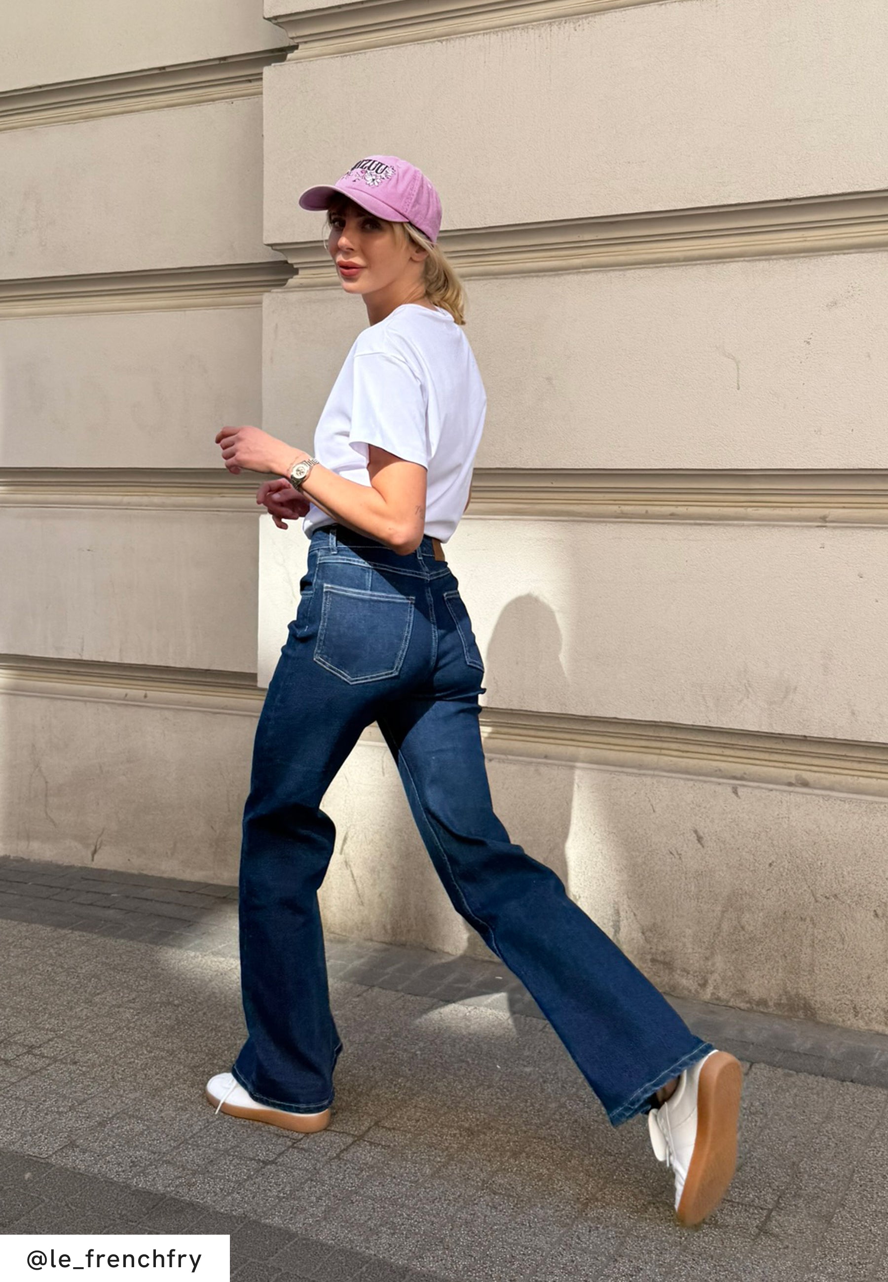 Ciemnoniebieskie jeansowe spodnie dzwony noszone z białym t-shirtem i białymi butami. Osoba idzie chodnikiem, ma na głowie różową czapkę z daszkiem.