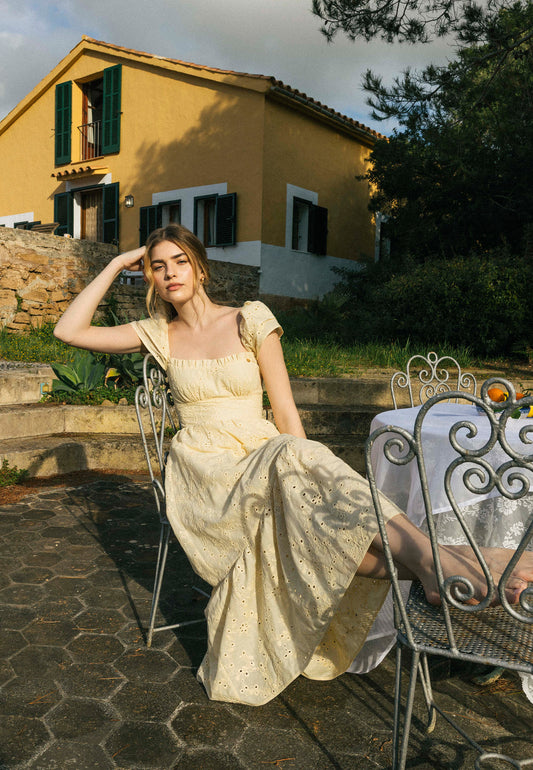 Modelka siedzi w ogrodzie przy stole, ubrana w żółtą sukienkę z koronki CORSICA.