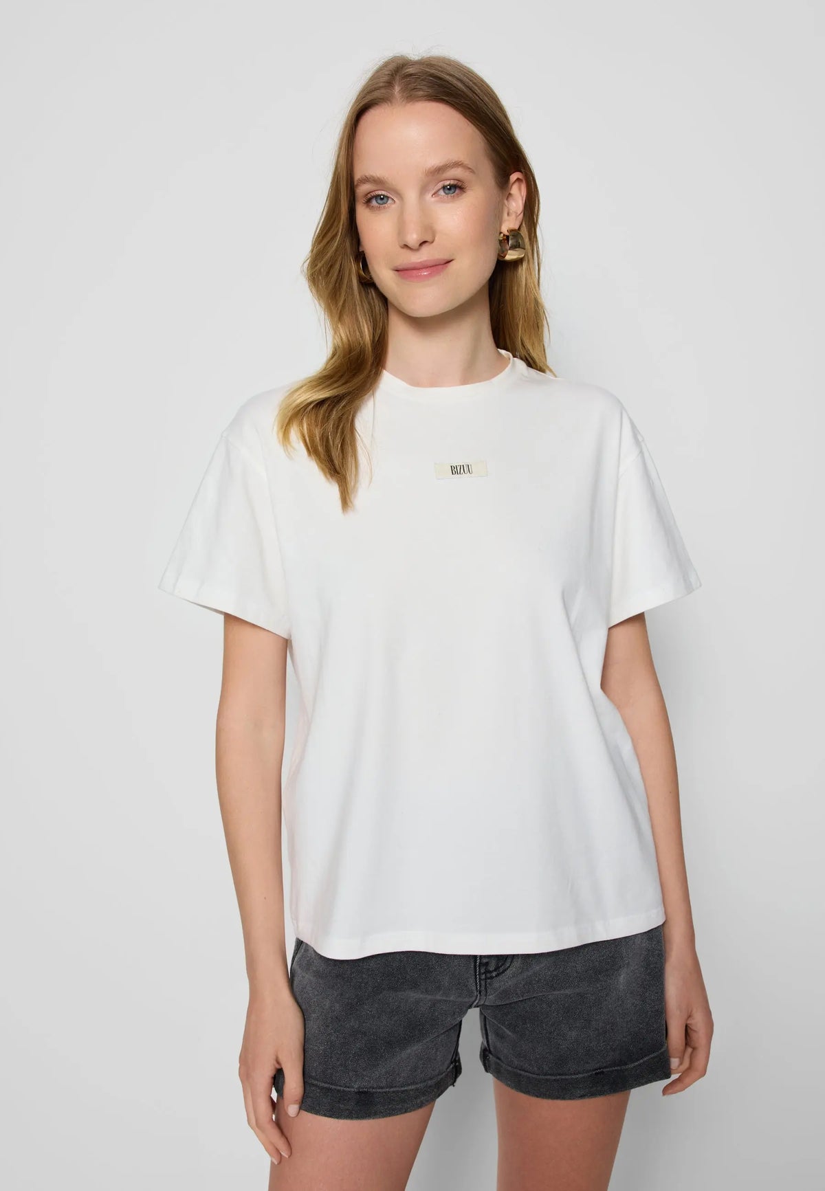 Modelka na białym tle w kremowym T-shircie TISHALLE i jeansowych szortach MENORCA.