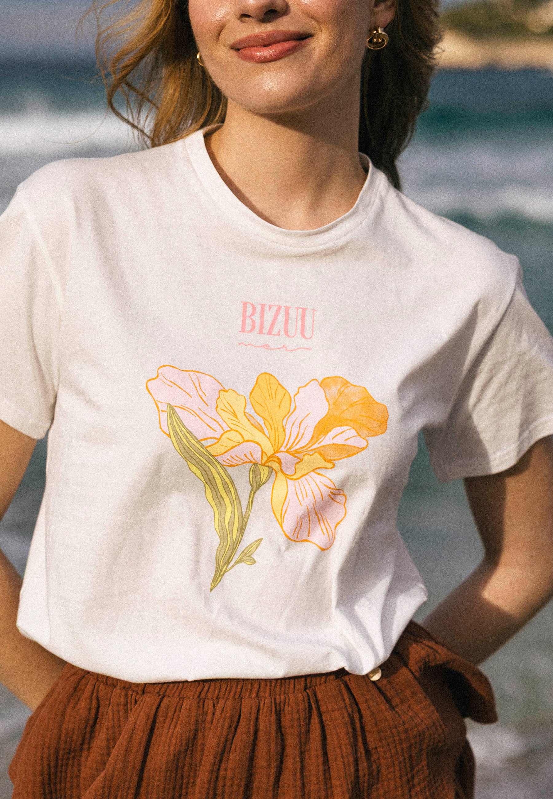 Zbliżenie na biały t-shirt TISAL z nadrukiem żółto-różowego kwiatu  i różowym napisem BIZUU, noszony do brązowych szortów OSTER.